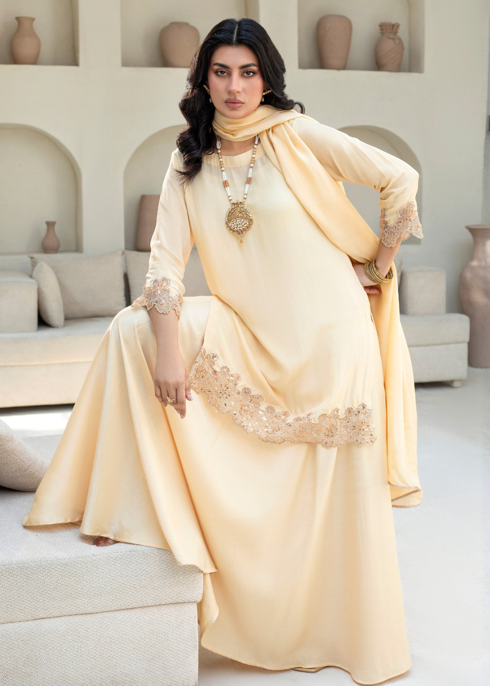 A woman models a light beige/cream traditional South Asian outfit consisting of a long kurta with lace trim, a matching dupatta, and a coordinating skirt, set in a modern, neutral-toned living room.