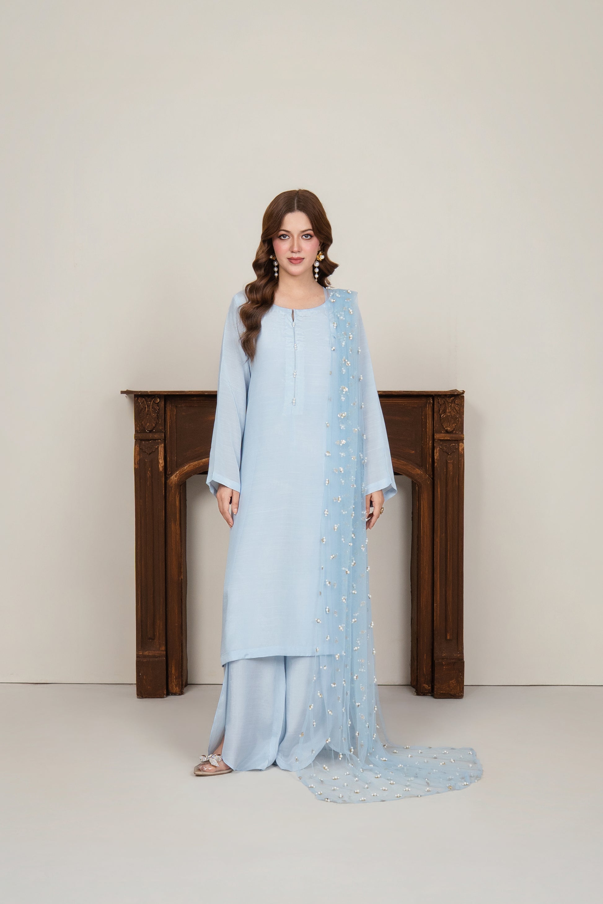 Full-length view of a woman wearing an ice blue raw silk kurta set featuring petal pants and a pearl-embellished neckline, paired with a matching net dupatta.